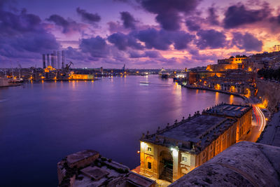 Illuminated buildings by sea against sky at sunset