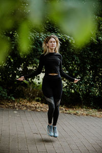 Full length of smiling young woman rope skipping under a tree