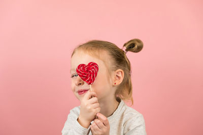 Cute girl holding heart shape on eye against colored background