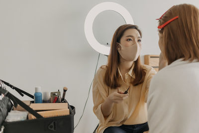 Side view of beautician wearing mask applying make up to woman