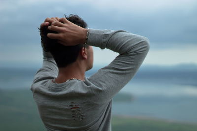 Rear view of man with hand in hair standing against sky