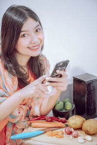 Portrait of smiling woman using phone while cutting vegetables at home
