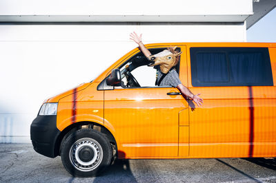 Man wearing horse mask sitting in orange van with arms outstretched on sunny day