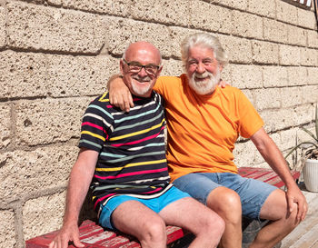 Portrait of smiling senior men sitting on bench against wall during sunny day