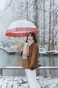 Portrait of young woman with umbrella standing by lake during winter