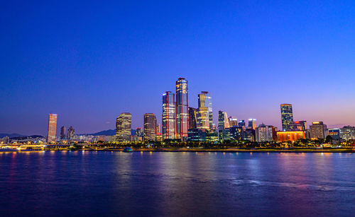 Illuminated modern buildings by bay against clear blue sky