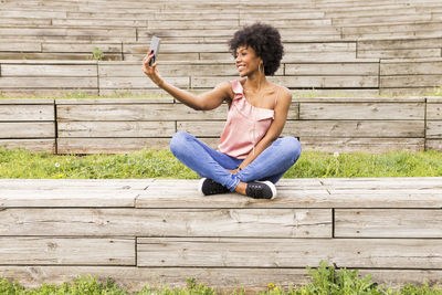 Smiling young woman taking selfie while sitting on wooden seat