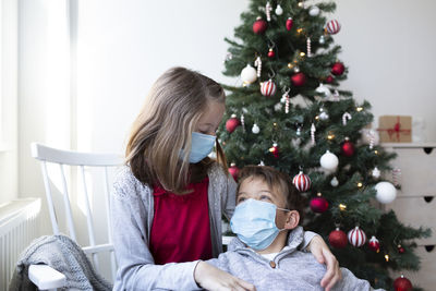 Siblings wearing mask against christmas tree at home