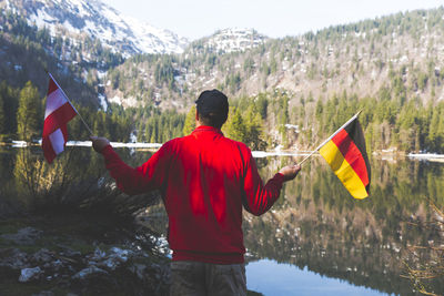 Rear view of mature man holding national flags while standing by lake against mountain