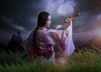 Rear view of young woman in traditional clothing holding sword while standing on field at night
