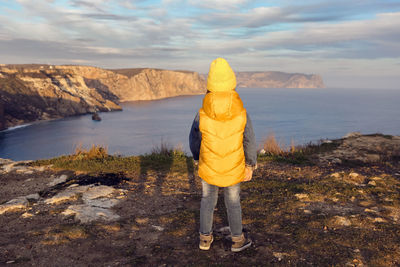 Boy child in a yellow hat and vest stands rear on top of a mountain by the sea in autumn