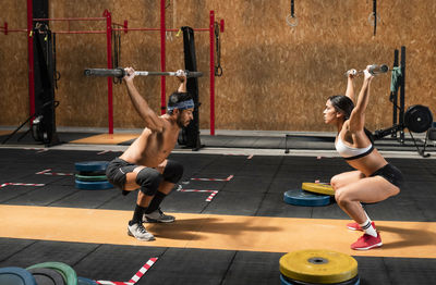 Side view of muscular sportsman and sportswoman doing squats with barbells while training actively in gym together and looking away