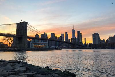 Bridge over river by buildings against sky during sunset