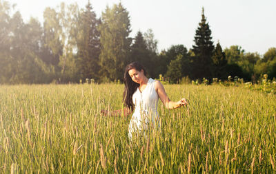 A young woman with long dark hair stands in a field with and looks at the ears of wheat. 