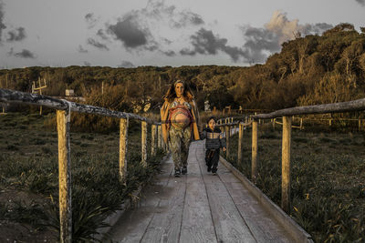 Woman walking with son on boardwalk against trees during sunset