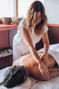 Female masseur with long hair in white uniform doing back massage to man lying on couch in bright salon room at daytime