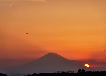 Scenic view of silhouette mountains and airplane flying against orange sky