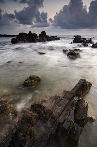 Sea waves splashing on rock formation against sky during sunset