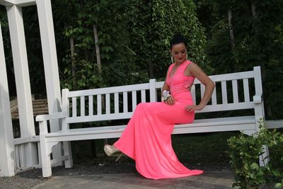 Full length of beautiful bride in wedding dress sitting on white bench in garden