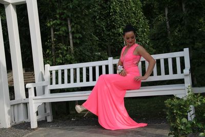 Full length of beautiful bride in wedding dress sitting on white bench in garden