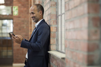 Side view of businessman using smart phone while standing against brick wall