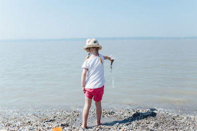 Girl wearing hat pouring water from container on shore at beach during sunny day