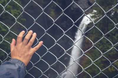 Cropped hand of person on fence against waterfall