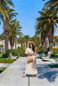 Full length portrait of beautiful woman wearing summer style dress on promenade under palm trees