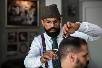 Serious mature bearded ethnic male barber trimming hair of client with scissors during grooming procedure in beauty salon