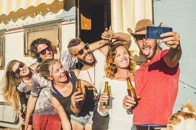 Friends holding beer bottles taking selfie standing by motor home