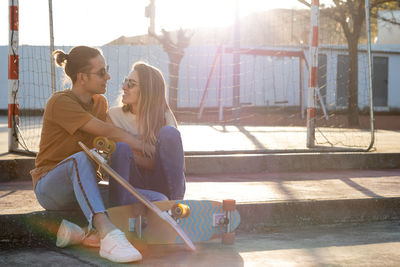 In love couple looking to each other sitting in park with two skate boards and sunset behind them