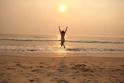 Rear view of woman jumping on shore at beach against sky during sunset