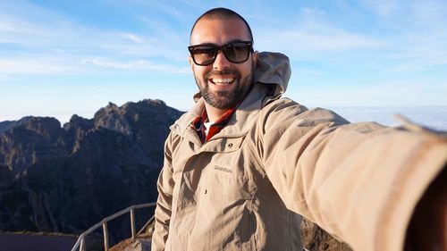 Smiling young man taking selfie while standing by mountain against sky