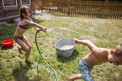 Playful sister spraying water on shirtless brother through hose at yard