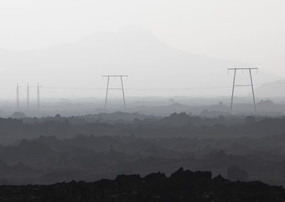 Black and white of power lines with wires placed on ground with rough surface against mountain range covered with fog