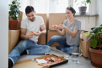 Smiling couple eating pizza while sitting against cardboard boxes in new house