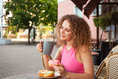 Young attractive curly woman sits at table in cafe on summer terrace. colorful delicious donuts, 