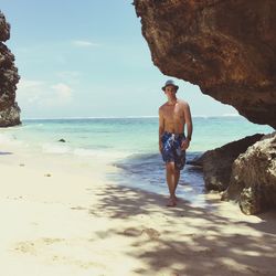 Portrait of shirtless young man walking by rock formation at beach