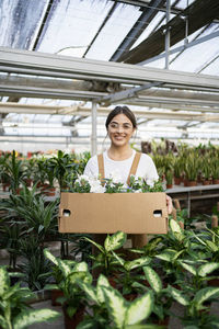 Happy gardener with box of flowering plants standing in greenhouse