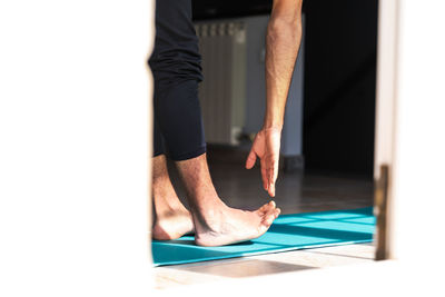 Side view of unrecognizable flexible barefoot man in sportswear doing standing forward bend exercise while training alone on sports mat at home