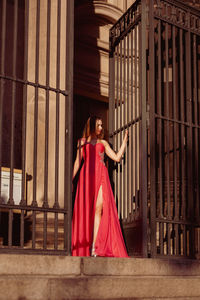 Full length of woman wearing red evening gown standing by metallic gate