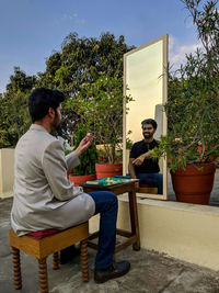 Young man sitting on potted plant against trees