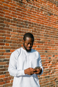 Young man using mobile phone while standing against brick wall