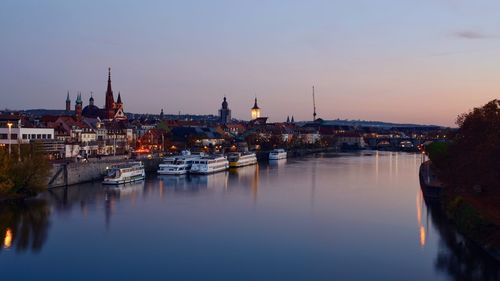 Sailboats moored on river by buildings against sky at dusk