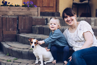 Portrait of smiling woman sitting by daughter playing with dog on steps