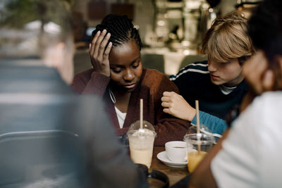 Teenage boy consoling female friend while sitting at cafe seen through glass window