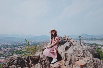 Full length portrait of woman sitting on rock against sky