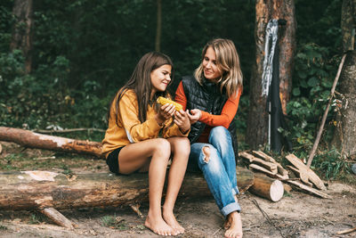A teenage girl with her mother is sitting on a log in the forest, a girl is eating corn