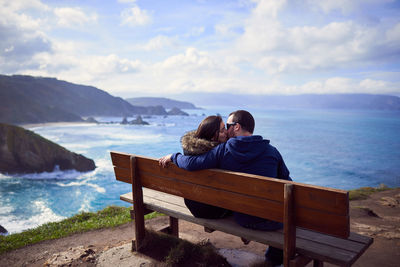 Rear view of couple embracing while sitting on bench by sea against sky