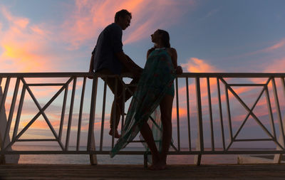 Low angle view of couple talking in balcony against cloudy sky during sunset
